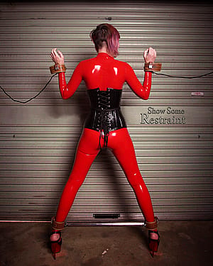 Foxxy in red latex catsuit, leather cuffs, spreader bar, and little black corset'